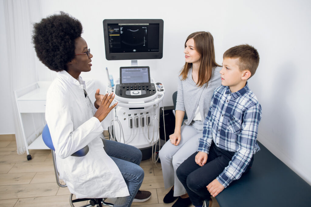 Young 30-aged Caucasian woman with her teen son visiting doctor at modern ultrasound exam room. Afro-american female doctor explaining procedure of ultrasound scan to her patients