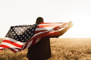 Young man holding American flag on back while standing in wheat field on dawn
