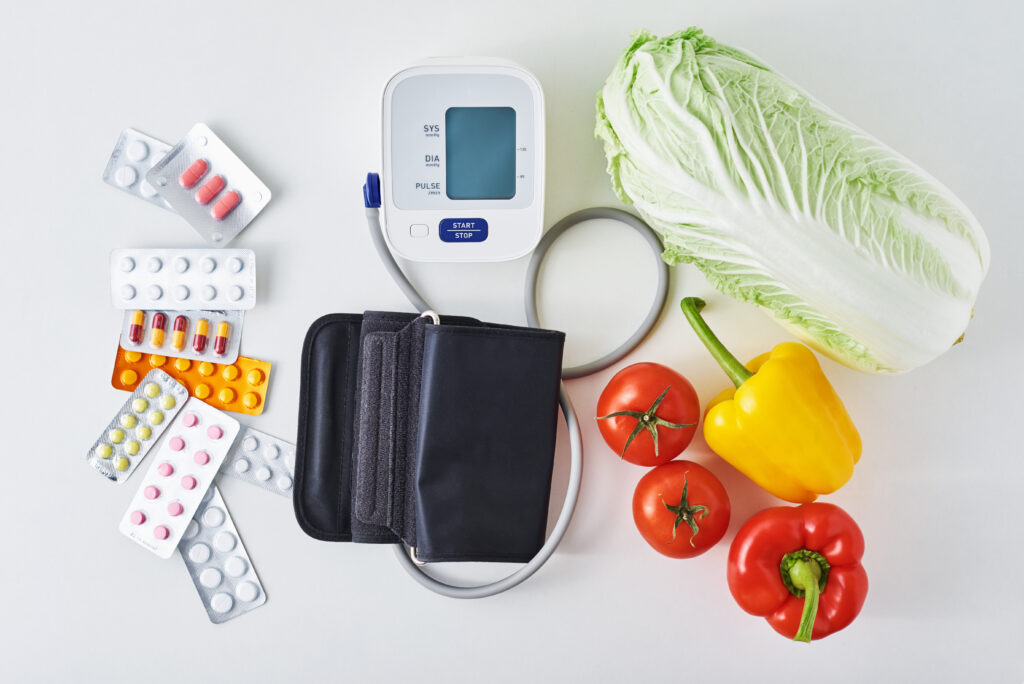 Digital blood pressure monitor, fresh vegetables and medical pills on white background. Healthy lifestyle concept