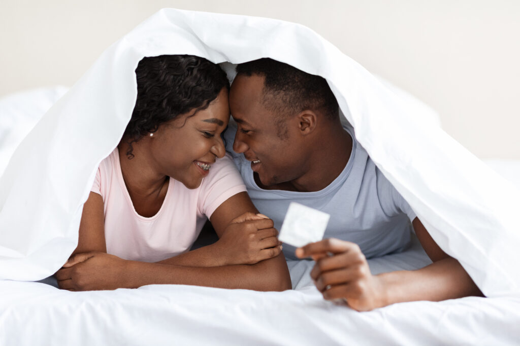 Romantic african american couple cuddling under blanket, man holding condom, copy space. Young black man and woman in love laying on bed at home, touching each other with foreheads and smiling