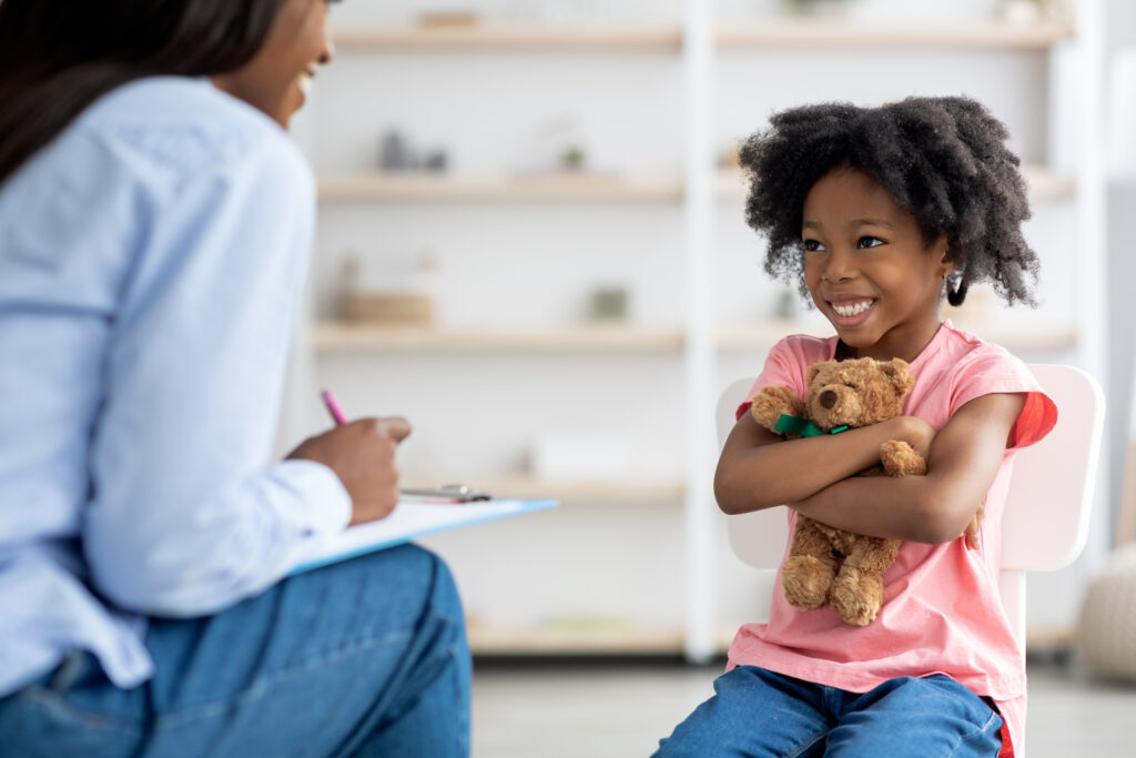 Happy little african american girl with cute curly hair sitting on chair at therapy session with child psychologist, hugging her teddy bear and smiling at unrecognizable black woman