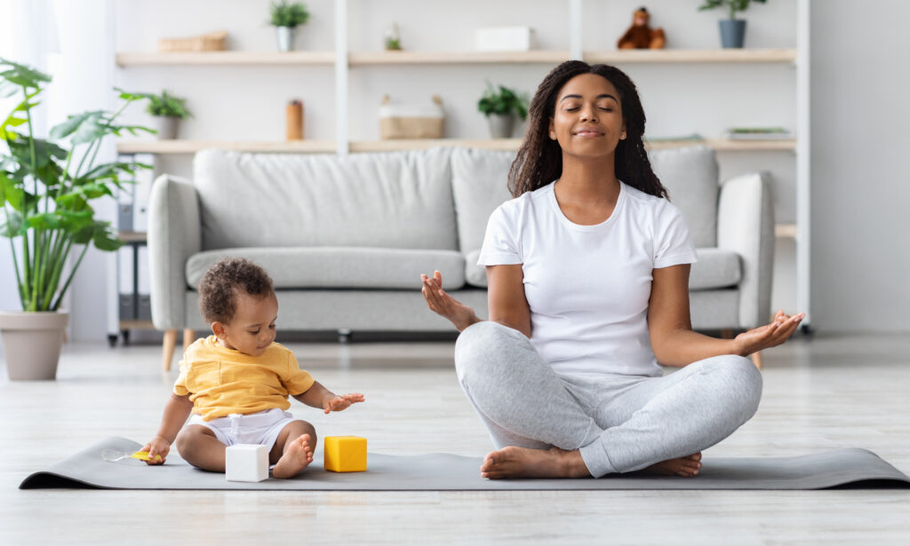 Calm Happy Black Mother Meditating With Baby At Home, African Lady Sitting In Lotus Position With Closed Eyes, Practicing Yoga In Living Room With Child