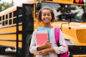Smiling African American schoolgirl going back to school with books waiting for school bus.