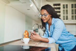 Young African woman leaning on her kitchen counter at home reading text messages on a cellphone and drinking a cup of coffee