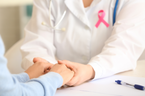 A doctor with a breast cancer awareness ribbon on their labcoat is holding the hands of an adult woman.